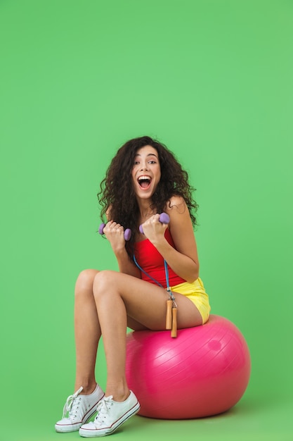 athletic woman wearing summer clothes lifting dumbbells while sitting on fitness ball during aerobics against green wall