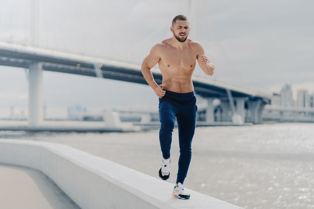 Il giovane atletico barbuto si dedica allo sport ha una corsa mattutina respira profondamente si prepara per la maratona lo sportivo motivato che fa jogging sul fiume ha un corpo forte e sano concetto di fitness e sport