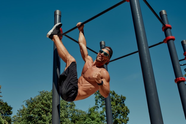 Athletic young man hanging from the bars at the calisthenics gym outdoors smiling