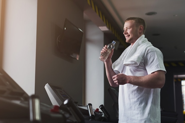 Athletic young man having rest after running training, drinking water at gym, standing at treadmill, copy space. Bodybuilding concept