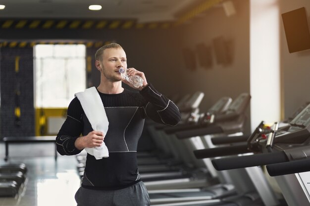 Athletic young man having rest after running training, drinking water at gym, standing at treadmill, copy space. Bodybuilding concept