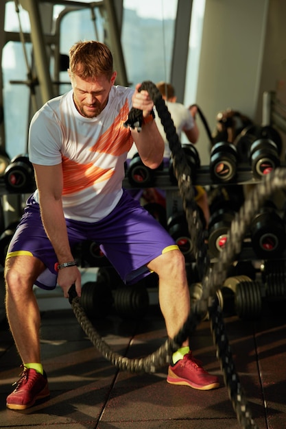 Athletic young man with battle rope doing exercise in functional training fitness gym