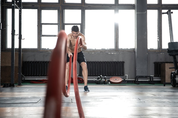 Athletic young man with battle rope doing exercise in functional training fitness gym