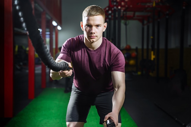 Athletic young man with battle rope doing exercise in functional training fitness gym