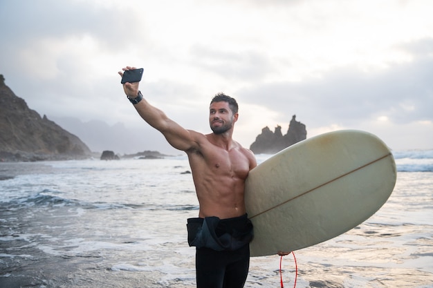 Athletic young surfer takes a selfie at the beach, sporty guy holds a surfboard and stands at a tropical beach during sunset - sporty millennial using technology and practicing extreme sport.