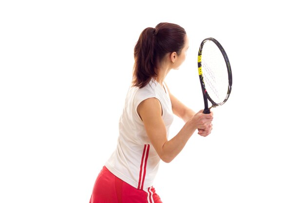 Athletic young woman in white sports shirt and red skirt with dark ponytail holding tennis raquet on white background in studio