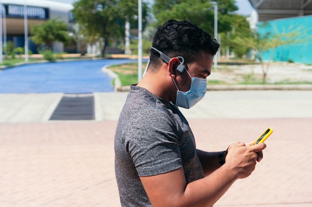 Atleet die een beschermend masker draagt en naar muziek luistert met een koptelefoon terwijl hij in het park staat en een mobiele telefoon vasthoudt