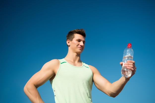 Atleet glimlachend in groene tshirt op blauwe hemel Bodybuilder met spieren biceps triceps op zonnige dag Gezonde levensstijl en gezondheid Sport en fitness concept Man met waterfles in gespierde handen