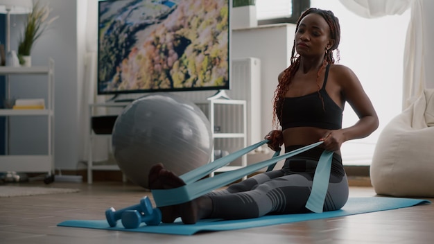 Atleet met zwarte huid in sportkleding die lichaamsspier uitoefent met behulp van fitnesselastiek en geniet van een gezonde levensstijl zittend op een yogakaart in de woonkamer. Fit vrouw die werkt bij wellness-opwarming voor de training