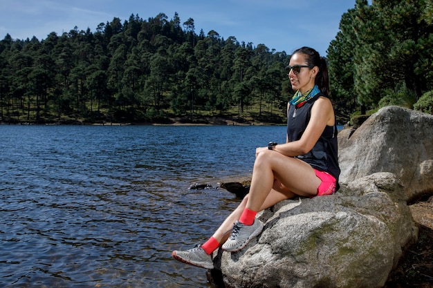 Atleta femenino descansando sobre een roca a la orilla de un lago despus de correr en el bosque