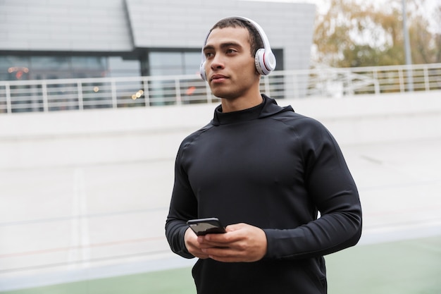 atletische blanke sportman in trainingspak met koptelefoon en mobiele telefoon tijdens het sporten in het stadion na regen