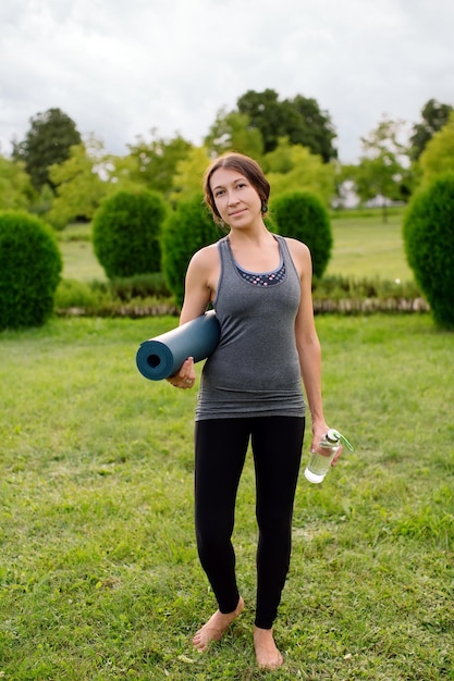 Atletische dunne jonge vrouw houdt yogamat en fles water vast na of voor de training in het buitenpark.