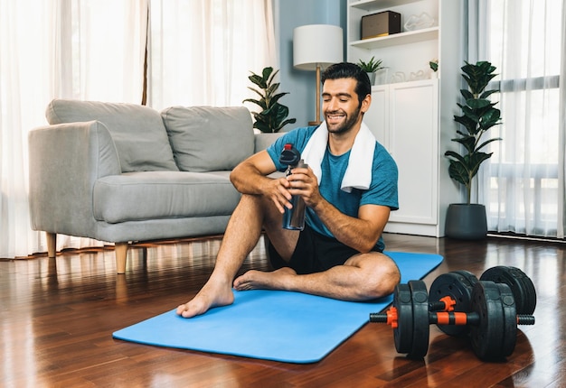 Atletische en sportieve man die water drinkt op een fitnessmat na het afmaken van een thuis workout sessie voor een fit lichaam en een gezonde sport levensstijl thuis Gaiety thuis workout training.