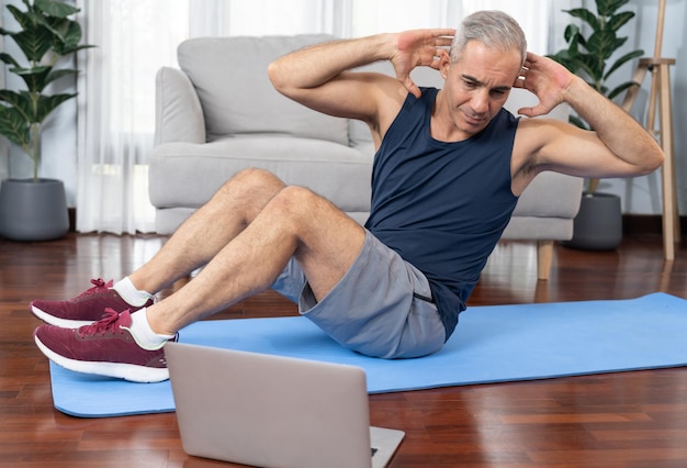 Atletische en sportieve senior man doet crunch op de oefenmat tijdens het kijken naar online trainingssessie thuis oefenen als concept van een gezonde, fitte lichaamslevensstijl na pensionering