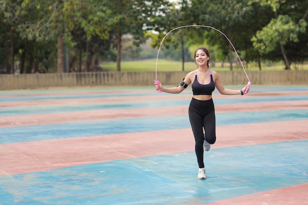 Atletische jonge Aziatische vrouw in sportkleding staande en springtouw buiten voor ochtendoefening. Actieve en gezonde meisjestraining op bicepsoefening met boomachtergrond. Sport en lifestyle concept