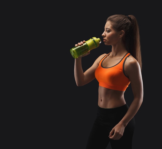 Atletische jonge vrouw die rust heeft na bobybuilding training, drinkwater op een zwarte achtergrond. Studio-opname, low key, kopieerruimte, zijaanzicht