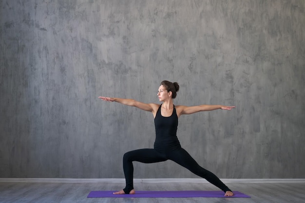 Atletische jonge vrouw die yoga doet die op grijze muurachtergrond in zwart wordt geïsoleerd