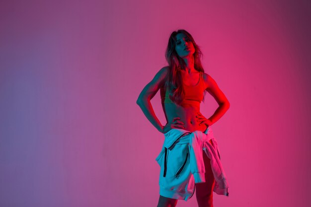 Atletische jonge vrouw in een vintage top in hoodie poseert in een kamer op een roze kleurrijke achtergrond. Sexy meisje mannequin in trendy slijtage staat in studio met fel neon roze licht.