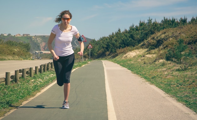 Atletische jonge vrouw met koptelefoon luisteren naar muziek van haar smartphone terwijl ze buiten op een landingsbaan rent. Modern gezond levensstijlconcept.