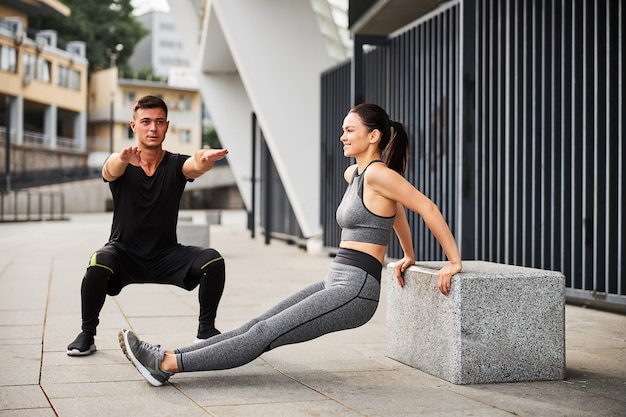 Atletische knappe man doet squats terwijl zijn vriendin buitenshuis push-ups doet voor triceps