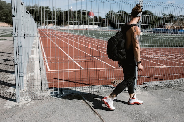 Atletische man in hoofdband gekleed in zwarte sportkleding met rugzak op zijn schouders loopt op een zonnige dag langs het hek van de speeltuin.