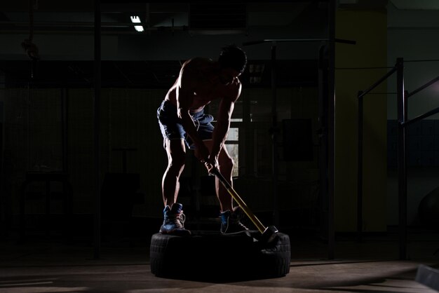 Atletische man slaat bandentraining in sportschool met hamer en tractorband Siluet