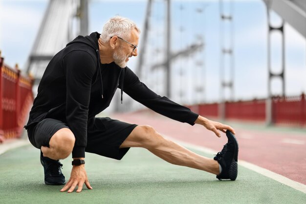 Atletische oudere sportman die zijn benen strekt, traint buiten op de loopbrug in de stad. Gemotiveerde, goed passende senior man in zwarte sportkleding aan het opwarmen, joggen op straat, kopieerruimte