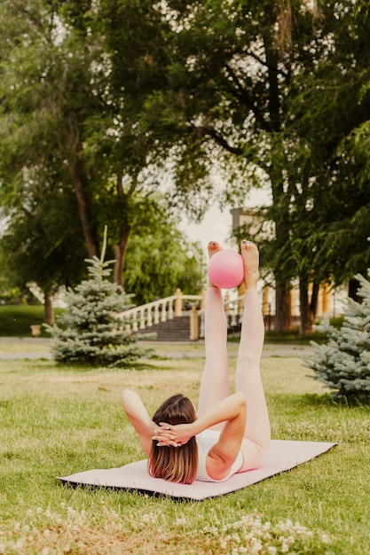 Atletische, slanke vrouw in roze kleding en kleine rubberen bal die Pilates doet op roze mat bij zonsopgang in het park in de zomer.