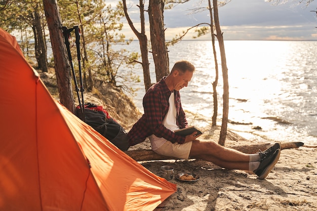 Atletische volwassen man gaat kamperen in het bos boven de kust en geniet van het ontbijt terwijl hij een boek leest