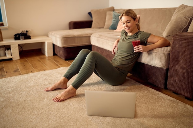 Atletische vrouw die smoothie drinkt terwijl ze laptop gebruikt en ontspant op de vloer