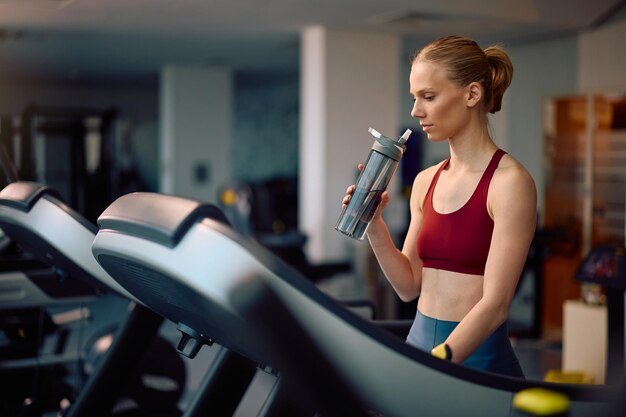 Atletische vrouw drinkt water terwijl ze oefent op een loopband in een sportschool
