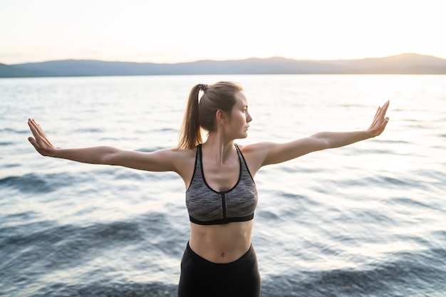 Atletische vrouw in een sport-bh en zwarte legging strekt zich uit voordat ze lessen begint aan de avondoever van het meer