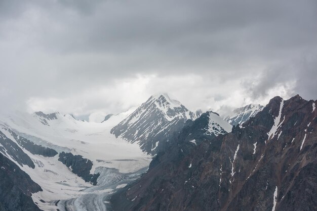 Foto atmosferisch landschap met scherpe rotsen en hoge besneeuwde bergtoppen in regenachtige lage wolken bij bewolking dramatisch somber landschap met grote sneeuwbergen en gletsjer in grijze bewolkte hemel bij regenachtig weer