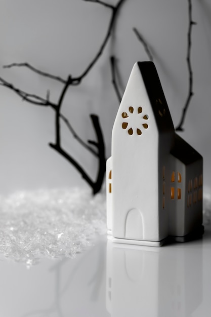 Photo atmospheric composition with white ceramic church candle house, snowflakes, tree branch on white wall.