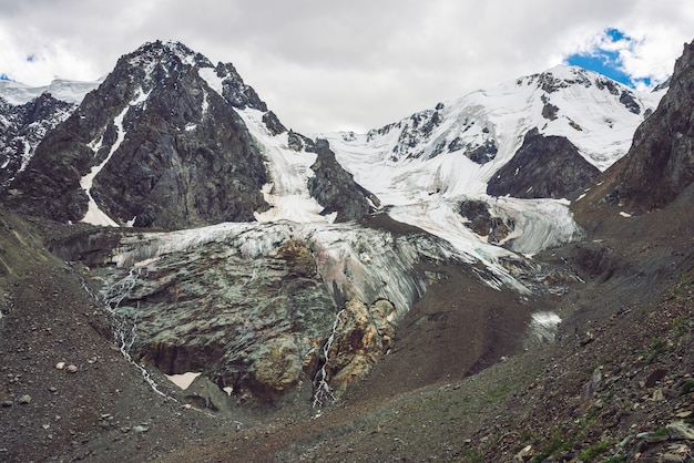 Atmospheric giant snowy mountains. Icy rocks with waterfall. Water stream from glacier. Dark rocky mountainside with ice. Snow on mountain ridge. Unusual landscape of majestic nature of highlands.