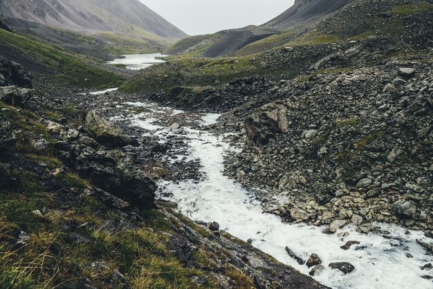 Atmospheric landscape with mountain lake and mountain creek among moraines in rainy weather. Bleak overcast scenery with milky river and lake among rocks. Gloomy view to milk mountain river and lake.