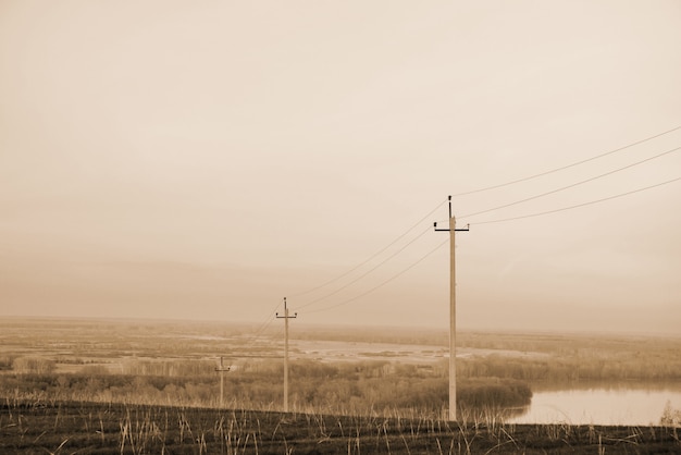 Atmospheric landscape with power lines in field on background of river under sepia sky.