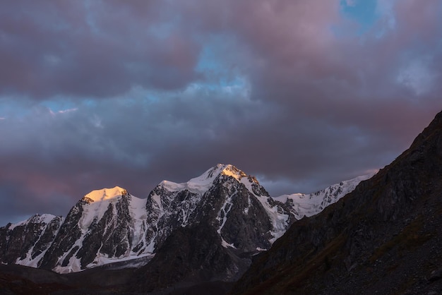 Atmospheric landscape with sunset gold reflection on huge snowy mountain top in violet dramatic sky Scenic view to giant snow mountains in dusk Snowcovered mountain range silhouettes in twilight