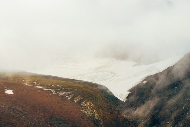 Атмосферный минималистский альпийский пейзаж в высокогорной долине с широким ледником на склоне горы внутри облака. Низкие облака в скалистой долине. Спокойный горный пейзаж. Прекрасный вид на снежные горы