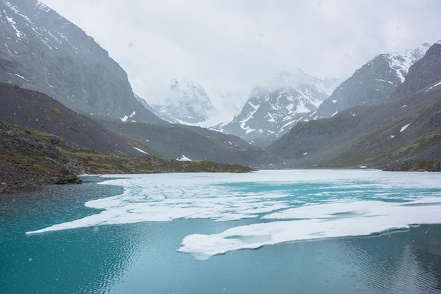 Atmosferico paesaggio montano con lago alpino ghiacciato durante la nevicata fantastico scenario freddo con nevicate in alta valle di montagna con lago di montagna ghiacciato sullo sfondo di montagne innevate tra nuvole basse