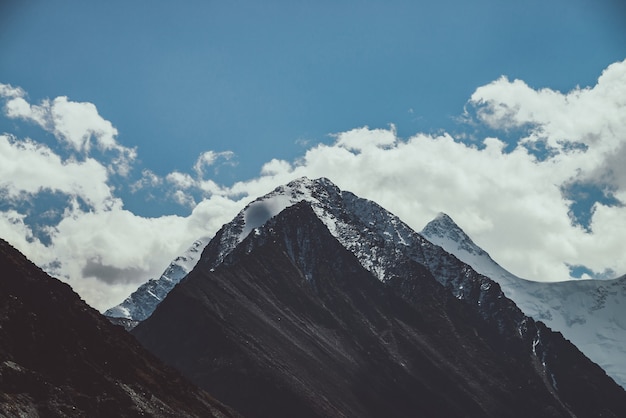 Atmospheric mountain landscape with great snow-covered pinnacle and snowy pointy peak in low clouds in dark colors. Gloomy mountain scenery with high snowy peaked top under cloudy sky in faded tones.