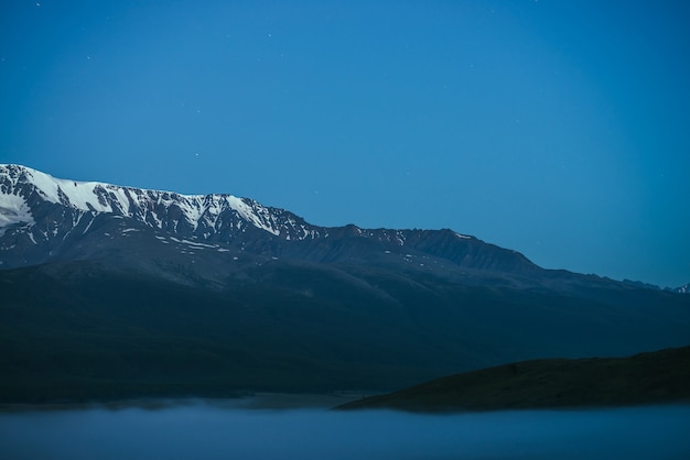 薄明かりの空の下で濃霧と素晴らしい雪の山脈のある大気中の山々の風景。夜の濃い霧の上に大きな雪山の尾根があるアルプスの風景。夕暮れ時に雲の上の雪の岩。