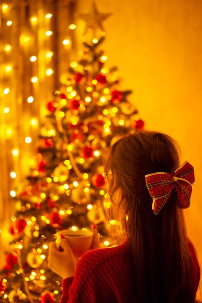 Atmospheric photo of a young girl admiring beauty of Christmas tree and decorations at home drinking hot tea Christmas festive atmosphere