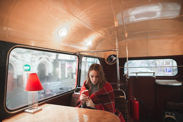 Atmospheric portrait of a girl sitting in coffee shop at the table, covered with a blanket