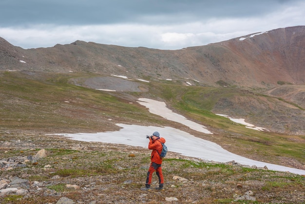 Atmospheric scenery with man in red against mountain range with sharp rocks under gray cloudy sky Tourist with camera shoots dramatic mountain landscape Man photographs mountains at gloomy weather