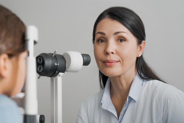 Attente optometrist die in de camera kijkt tijdens het onderzoeken van kindpatiënt op spleetlamp in kliniek