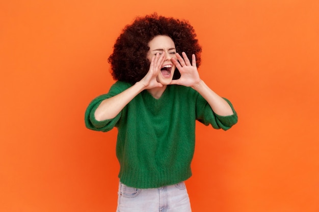 Photo attention announcement side view portrait of woman with afro hairstyle wearing green casual style sweater screaming message loud advertising indoor studio shot isolated on orange background