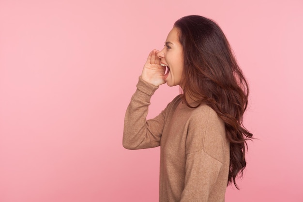 Attention, important message. Side view of young woman with brunette wavy hair shouting news or proclaiming idea with hand near mouth, announcing advertisement. studio shot isolated on pink background