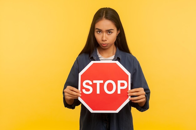 Foto attenzione, restrizioni. modo vietato. ritratto di ragazza in camicia di jeans con in mano rosso stop segnale stradale e guardando con espressione negativa di avvertimento. studio indoor girato isolato su sfondo giallo