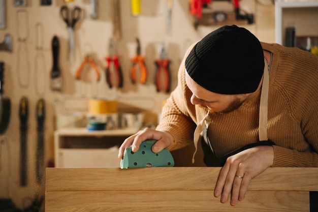 Attentive carpenter in a watch cap grinding a piece of wood,\
blurred background. in a big workshop. number of tools hanging on\
the wall.